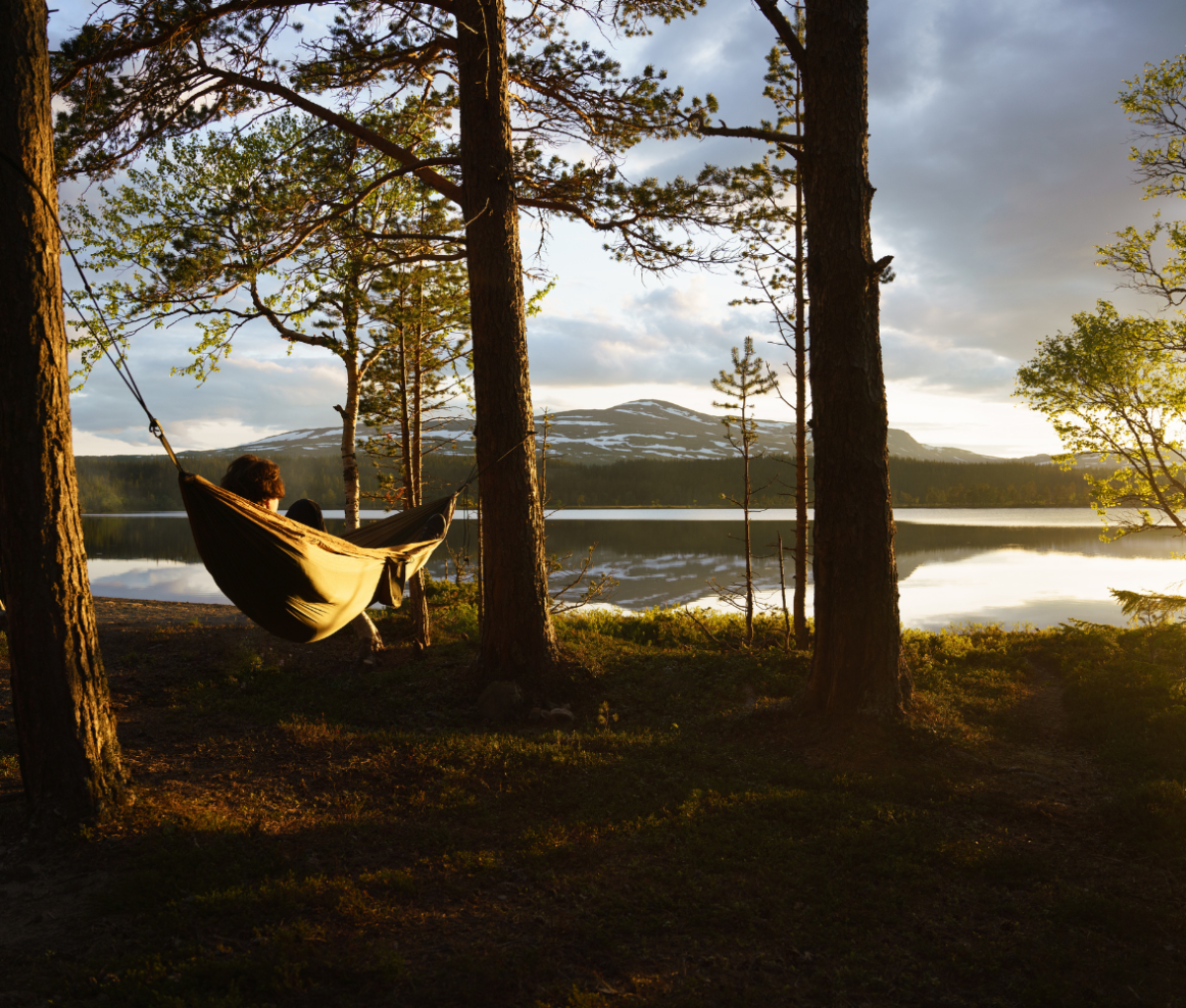 a person lying on a hammock amongst some trees by the side of a mountain lake in Jämtland Härjedalen, Sweden